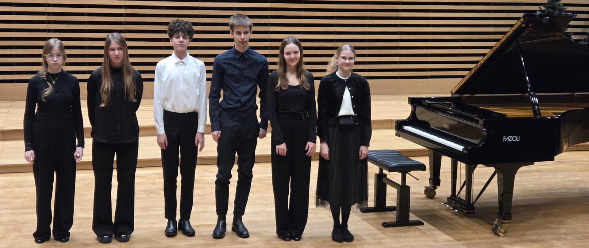 Wszyscy wykonawcy stoją na scenie przy fortepianie.