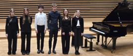 Wszyscy wykonawcy stoją na scenie przy fortepianie.