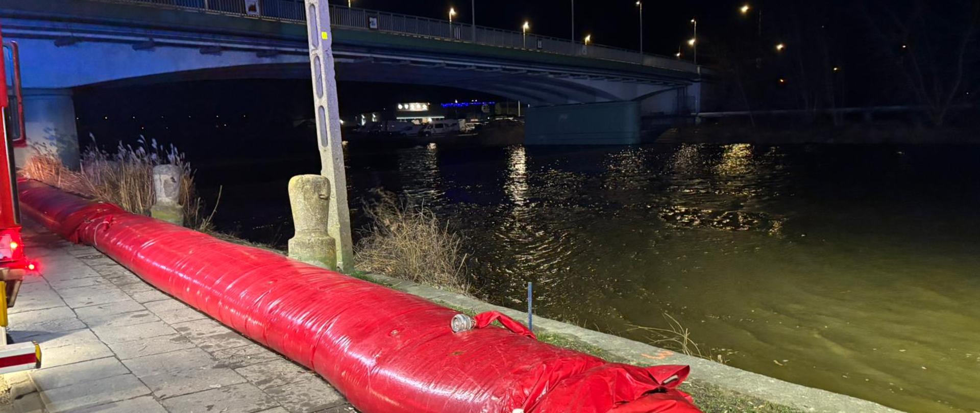 Obok rzeki, na jej brzegu znajduje się czerwony rękaw przeciwpowodziowy, w tle widać most.