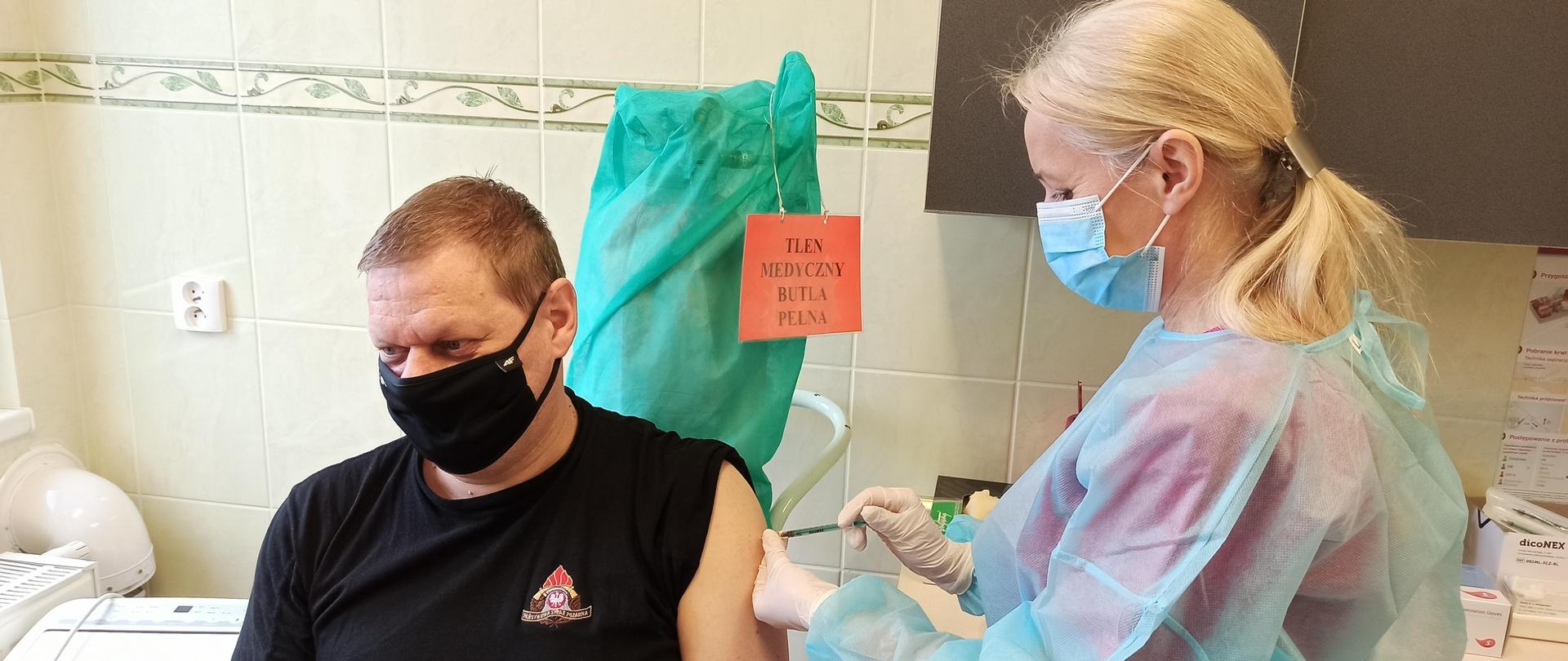 Zdjęcie przedstawia funkcjonariusza przyjmującego szczepionkę. Na pierwszym planie siedzący w gabinecie medycznym strażak z maseczką na twarzy. Pielęgniarka wstrzykuje szczepionkę strażakowi.