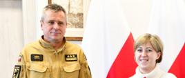 Pomorski komendant wojewódzki Państwowej Straży Pożarnej wspólnie z pracownikiem służby cywilnej stoją na tle flag. Pracownik trzyma czerwoną teczkę w ręce. 