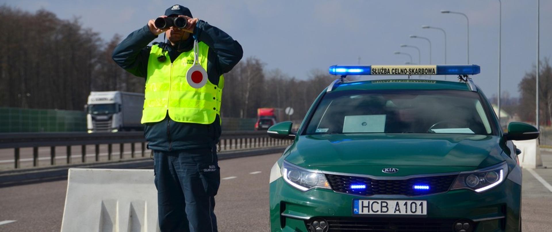 Funkcjonariusz patrzy przez lornetkę przy radiowozie