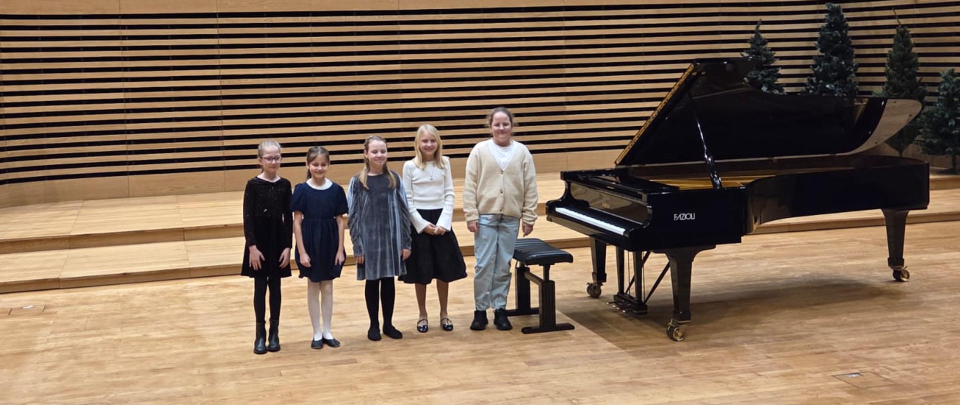 Pięć dziewczynek stoi na scenie przy fortepianie.