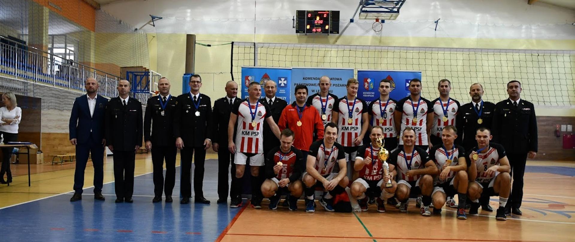 Na zdjęciu widoczni zawodnicy zwycięskiej drużyny z zawieszonymi na szyjach złotymi medalami w towarzystwie komendantów z rejonu województwa podkarpackiego w mundurach wyjściowych strażaka. Zawodnicy ubrani są w koszulki sportowe z napisem KM PSP w biało-czerwonych barwach. Jeden z zawodników znajdujący się w pierwszym rzędzie trzyma puchar za 1 miejsce.
