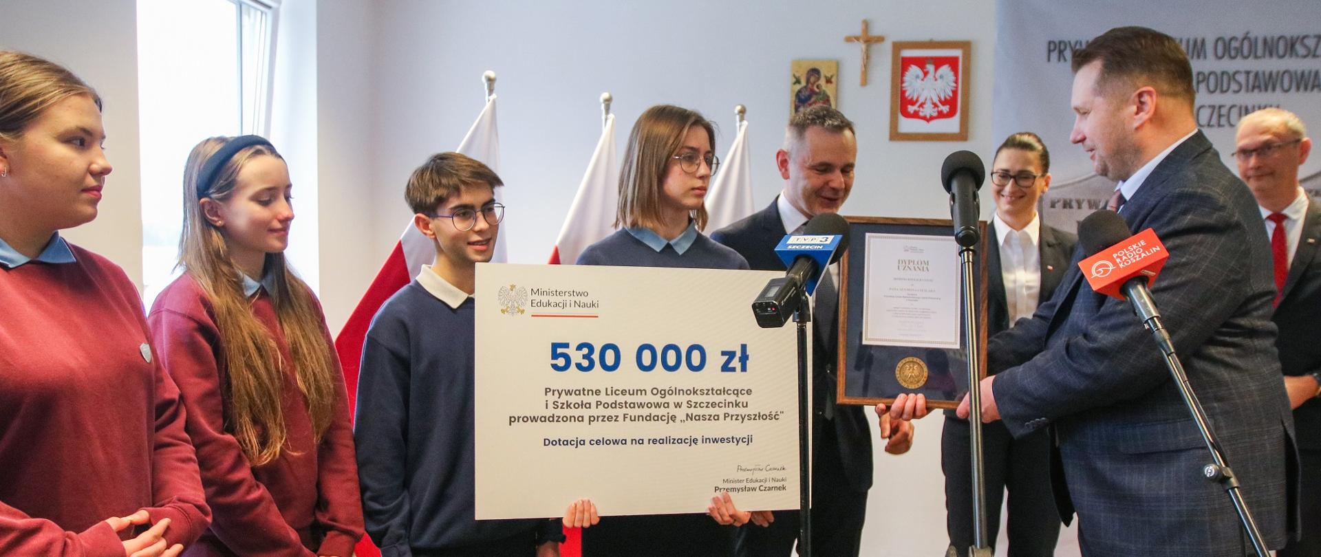 Minister Czarnek stoi obok grupy młodzieży w szarych i czerwonych bluzach, jedna dziewczyna trzyma wielki symboliczny czek z napisem 530 000 zł, przed nimi kilka mikrofonów na stojakach, w tle grupa dorosłych i polskie flagi.
