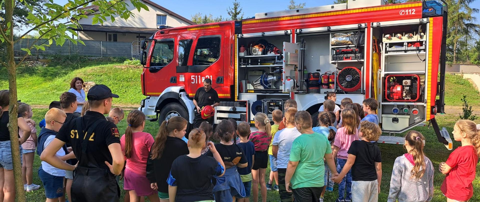Dzieci stoją tyłem na trawie na tle samochodu strażackiego, który ma pootwierane skrytki ze sprzętem.
