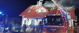 Na zdjęciu widać parterowy dom, w którym doszło do pożaru. Z dachu wydobywa się dym. Przed domem stoi podnośnik strażacki z wysuniętym koszem na dachem budynku. Podnośnik obsługuje strażak w żółtym stroju i czerwonym hełmem. Przed domem widać trzech strażaków w ciemnych strojach i białych hełmach. Przed ogrodzeniem posesji widać częściowo drugiego samochodu strażackiego. Jest ciemno.