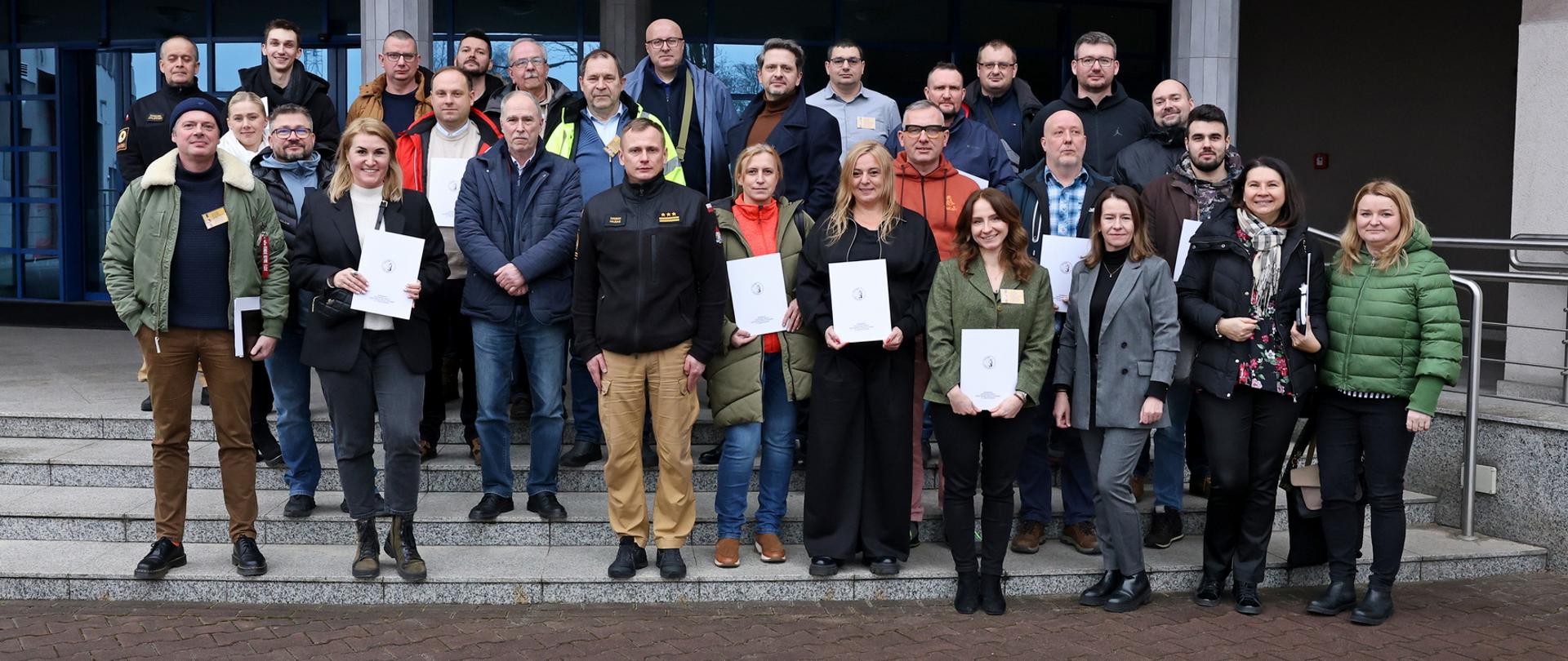 Pamiątkowe zdjęcie grupowe uczestników szkolenia inspektorów ochrony przeciwpożarowej (26 osób) w towarzystwie Zastępcy Komendanta Centralnej Szkoły PSP, kobiety oraz funkcjonariusza CS PSP