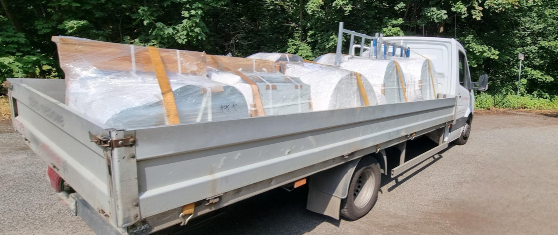 Na betonowym podłożu znajduje się zaparkowany biały pojazd dostawczy z widoczną przestrzenią ładunkową. Ładunek znajdujący się na pojeździe zabezpieczono pomarańczowymi pasami. Wokół znajdują się gęste drzewa i inna roślinność. Jest słoneczny dzień.