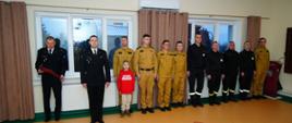 Fotografia przedstawia grupę strażaków i funkcjonariuszy ustawionych w szeregu podczas uroczystości. Wszyscy stoją w postawie wyprostowanej, co podkreśla oficjalny charakter wydarzenia. Wśród uczestników widoczne są osoby w różnych rodzajach umundurowania służbowego. Pomieszczenie jest dobrze oświetlone i przygotowane na ceremonię. Zdjęcie dokumentuje wspólny udział w ważnym wydarzeniu służbowym.