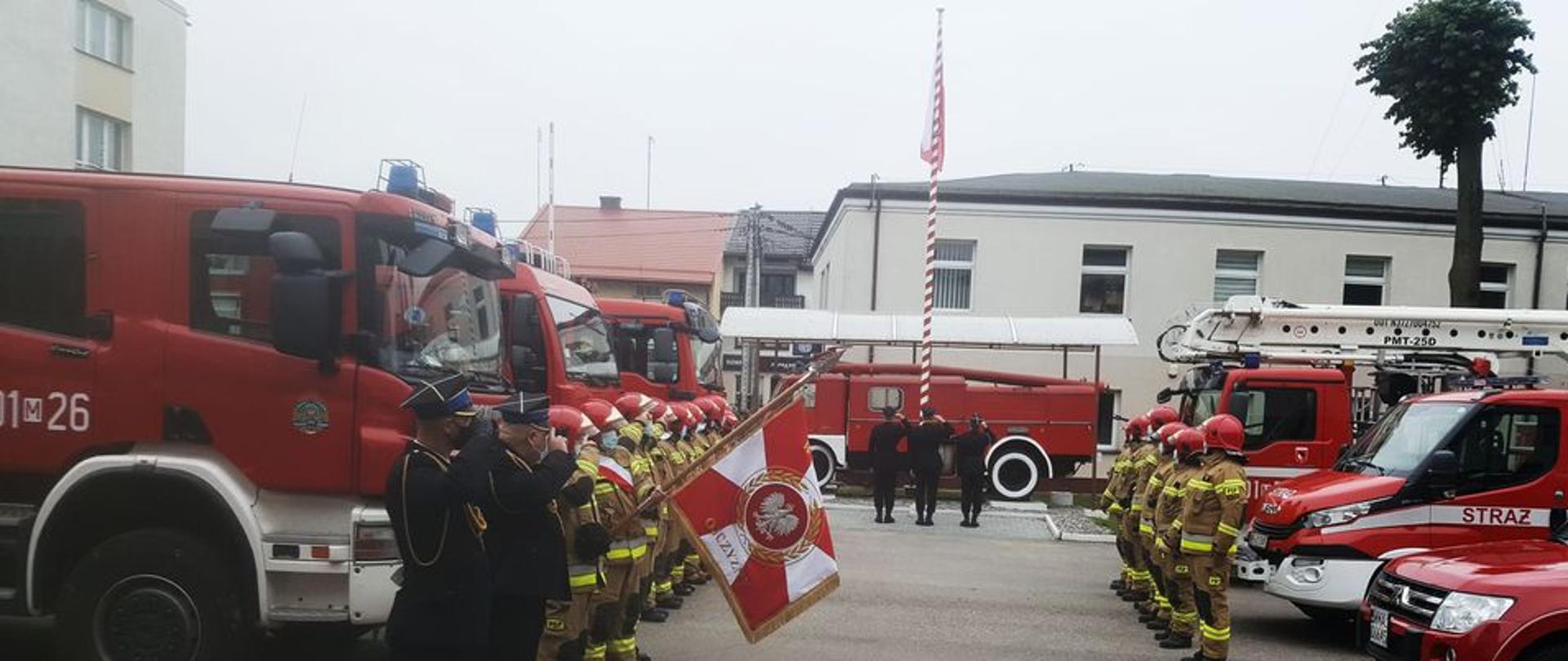 Trzech funkcjonariuszy w mundurach wyjściowych stoi przed flagą podniesioną na maszt. Funkcjonariusze w ubraniach specjalnych stoją naprzeciwko siebie przed samochodami pożarniczymi podczas uroczystej zmiany służby na placu wewnętrznym KP PSP. Po jednej stronie stoi także poczet sztandarowy w ubraniach wyjściowych. Poczet sztandarowy oraz poczet flagowy salutują.