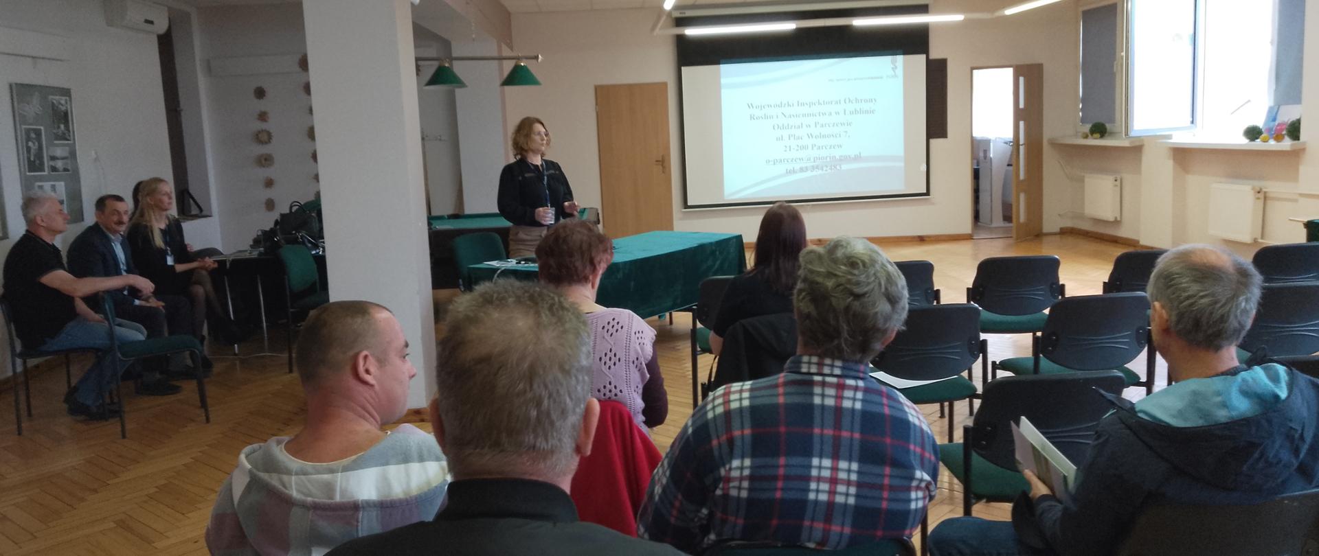 Na tle tablicy, na której wyświetla się slajd z nazwą Wojewódzki Inspektorat Ochrony Roślin i Nasiennictwa w Lublinie stoi kobieta. Na przeciwko kobiety siedzą rolnicy słuchający prelekcji.