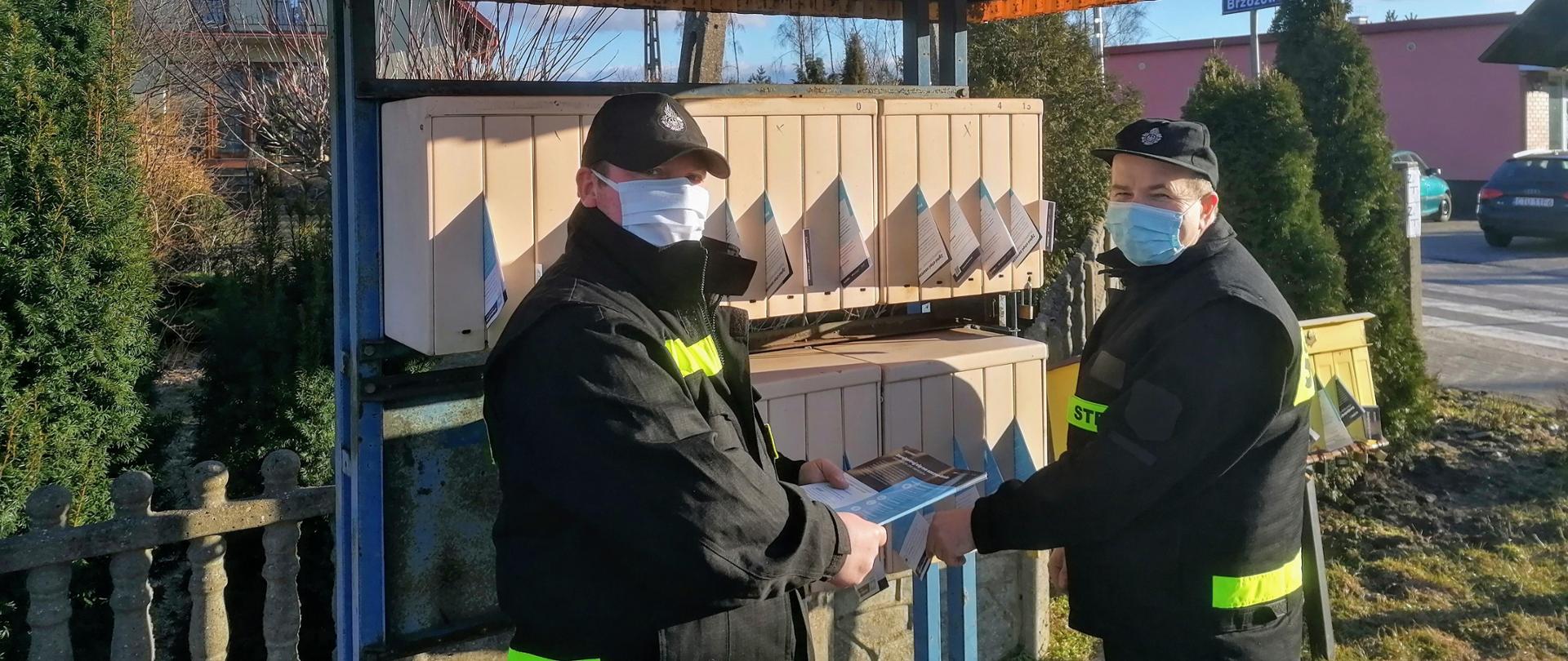 Zdjęcie przedstawia strażaków OSP w umundurowaniu koszarowym roznoszących ulotki na tle skrzynek na listy.