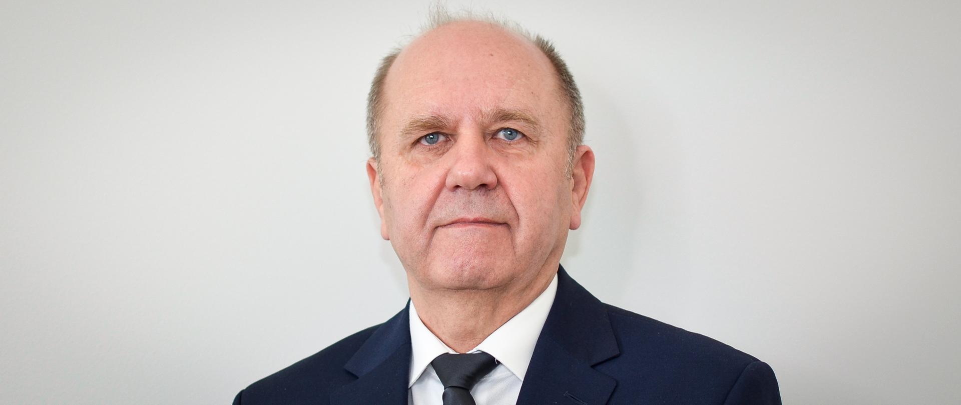 Mężczyzna w ciemnej marynarce, białej koszuli i ciemnym krawacie, z krótko przyciętymi włosami.