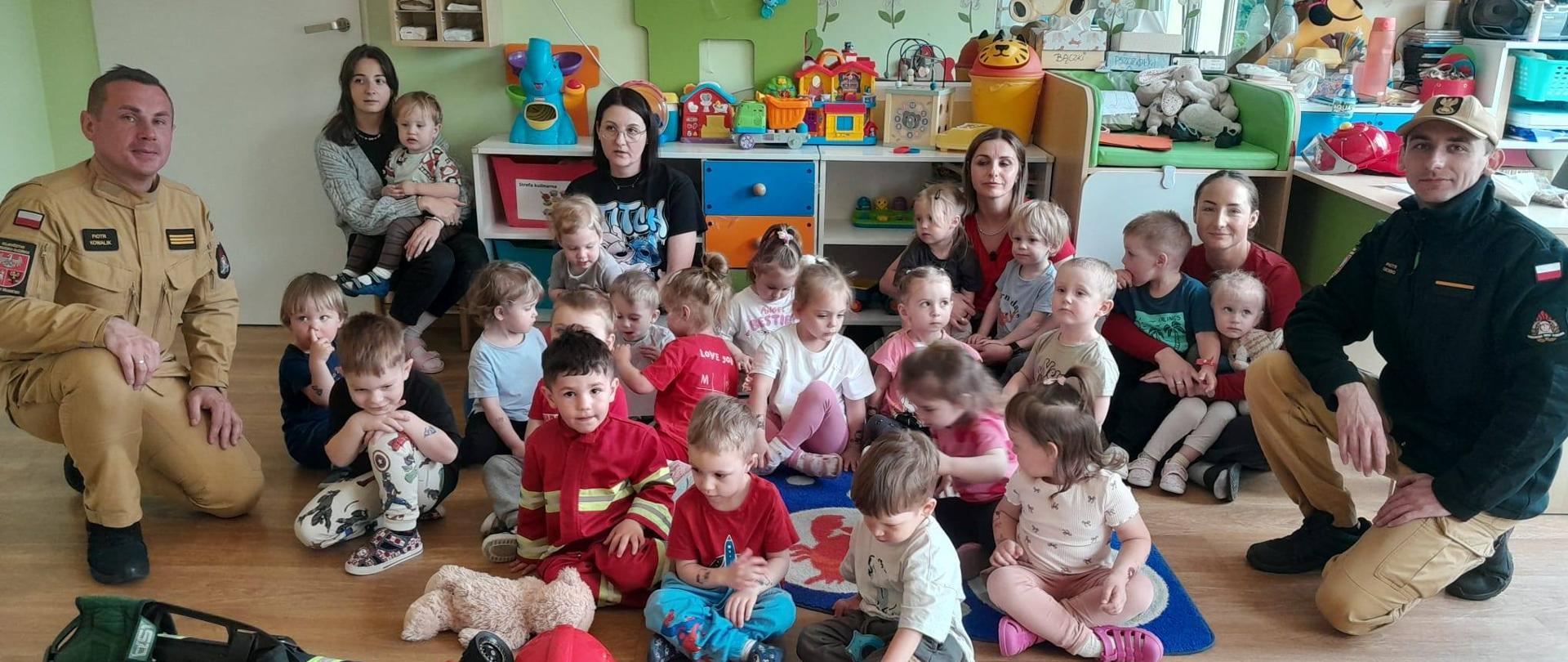 Sala przedszkolna, dzieci siedzą na podłodze z opiekunkami. Z prawej i lewej strony strażacy. W tle kolorowe mebelki i zabawki.