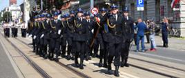 na zdjęciu pododdział honorowy Państwowej Straży Pożarnej maszerujący w trakcie defilady, na czele dowódca pododdziału, za nim sztandar komendy wojewódzkiego PSP w Łodzi i pododdział złożony z 20 strażaków, wszyscy w mundurach wyjściowych.