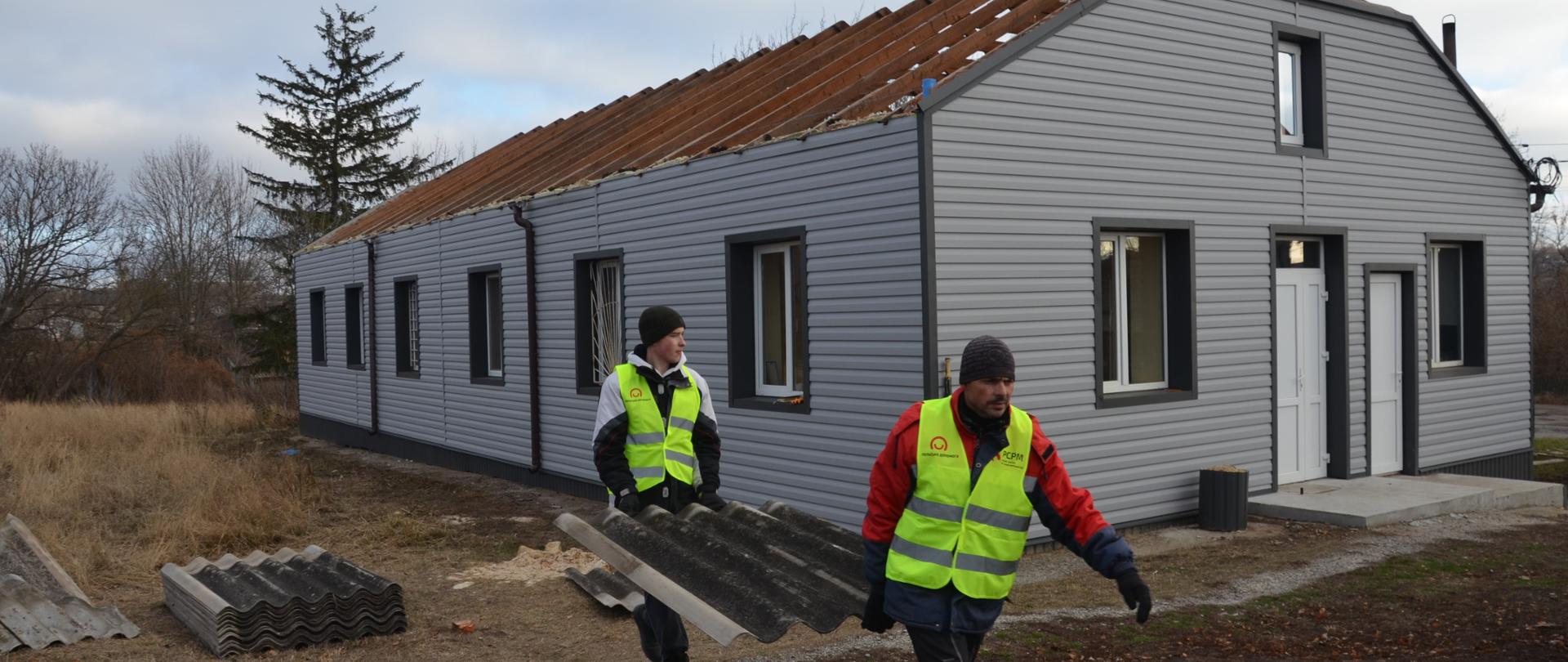 Dwóch mężczyzn niesie fragment dachówki. Ubrani są w kamizelki z logo Polska pomoc