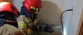 Dwóch strażaków ubranych w odzież ochrony osobistej klęczy na podłodze. Jeden z nich wykonuje otwór w ścianie przy pomocy wiertarki udarowej w celu dostania się do uwięzionego psa w przewodzie wentylacyjnym.