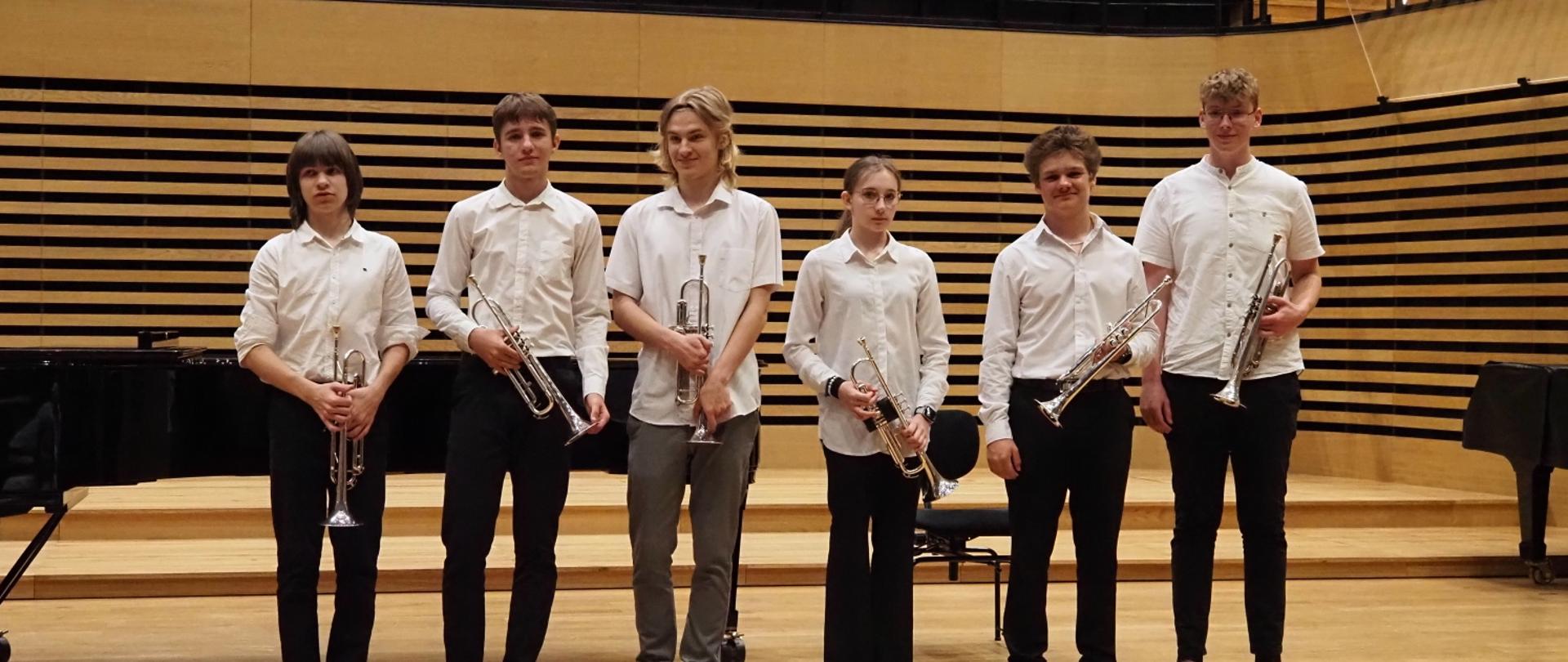 Na scenie stoi dziewczyna i pięciu chłopaków, wszyscy trzymają w rękach trąbki.