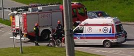 Samochód pożarniczy i karetka Pogotowia ratunkowego na wjeździe do Komendy. Na chodniku stoi rower, obok stoją strażacy.