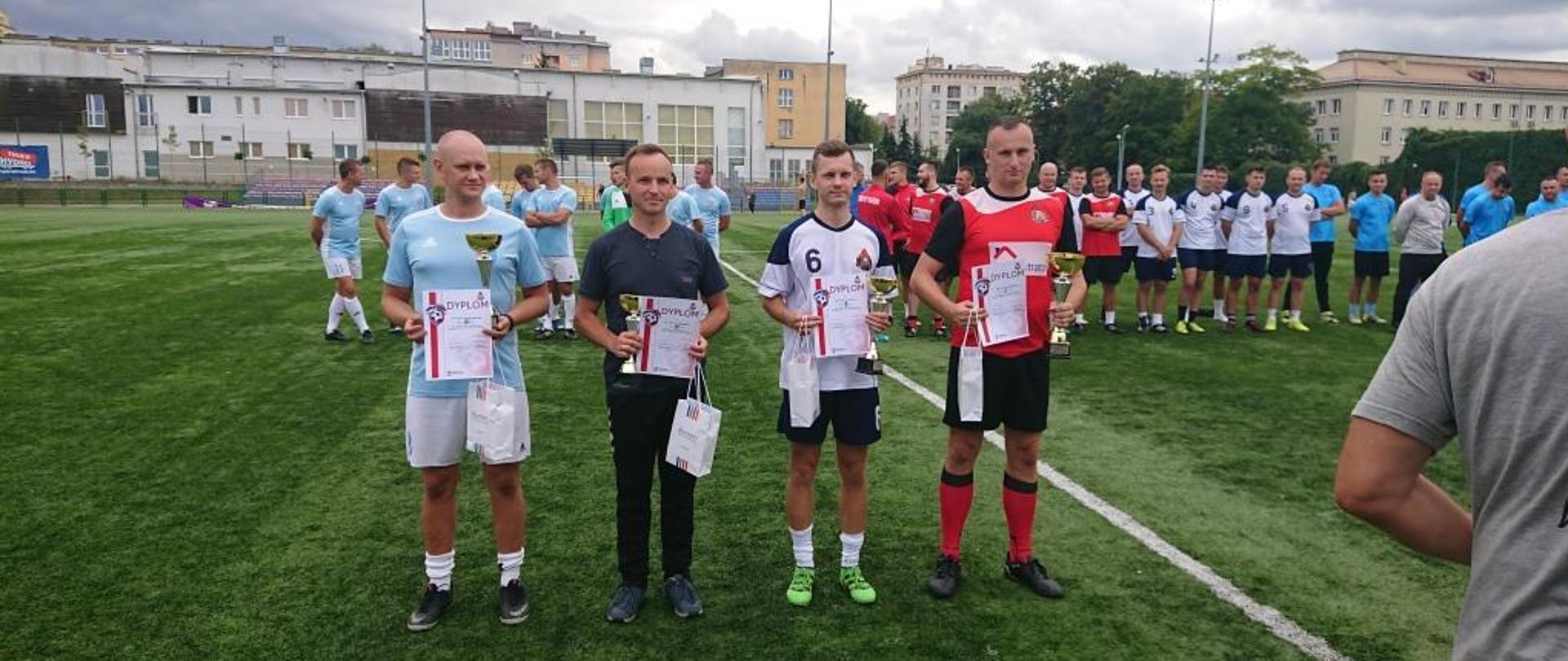 Zdjęcie wykonane na terenie Miejskiego Ośrodka Sportu i Rekreacji w Radomiu. Po ceremonii wręczenia pamiątkowych pucharów oraz dyplomów. Przed drużynami ustawieni są w szeregu ich kapitanowie. W dłoniach trzymają pamiątkowe upominki. Reprezentanci ubrani są w różnokolorowe stroje piłkarskie. 
