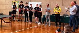 nauczyciele i uczniowie szkoły występują na sali gimnastycznej - Turniej Tenisa Stołowego Pamięci Mirka Czarnockiego 
