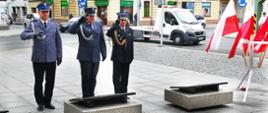 Upamiętnienie rocznicy. Plac przed pomnikiem Żołnierza Polskiego w Rawiczu. Delegacja rawickich służb mundurowych składa wiązankę kwiatów. Funkcjonariusze Policji, Służby Więziennej Państwowej Straży Pożarnej stoją przed pomnikiem w postawie zasadniczej i oddają honor poprzez salutowanie. Z prawej strony stojak z flagami państwowymi. W tle budynki, przechodnie i ruch uliczny.