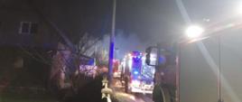 z lewej strony zdjęcia kawałek domku jednorodzinnego, za nim w oddali unoszą się kłęby dymu. Z prawej strony zdjęcia kolejno jeden za drugim ustawione trzy pojazdy pożarnicze z włączoną sygnalizacją świetlną.