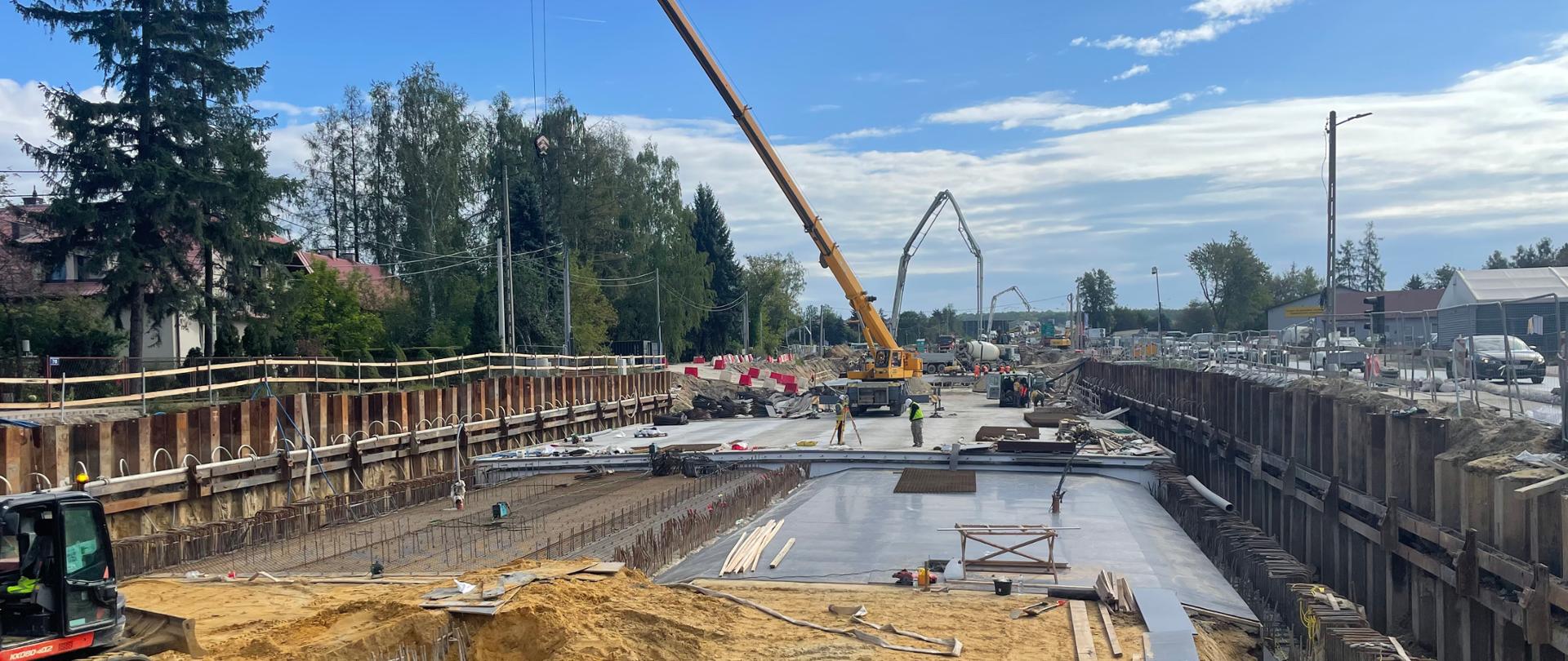 Zdjęcie przedstawia roboty drogowe. Na pierwszym planie widać utwardzanie gleby oraz żółty dźwig. Po bokach zabudowy domowe.