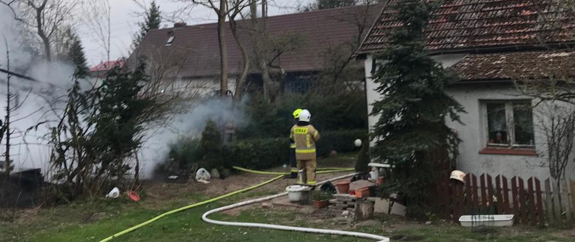 Zdjęcie przedstawia do jednorodzinny obok którego spłonęła stodoła. Na zdjęciu znajduje się dwóch strażaków oraz węże.