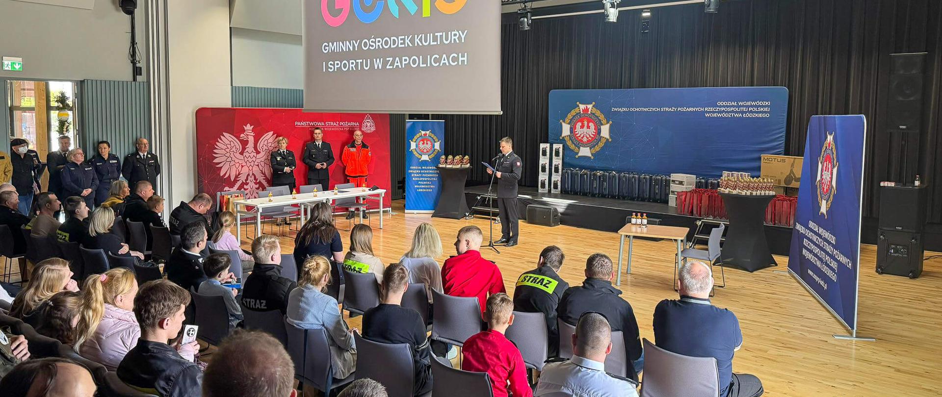 Widok na salę w Gminnym Ośrodku Kultury i Sportu w Zapolicach. Na scenie strażak przemawia do zgromadzonej publiczności. Nad sceną widoczny jest duży ekran z kolorowym logo ośrodka „GOKiS”.
