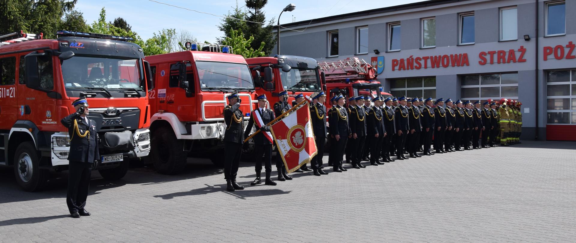 Dowódca wraz z pododdziałem i pocztami stoją na baczność podczas śpiewana hymnu narodowego, w tle samochody pożarnicze