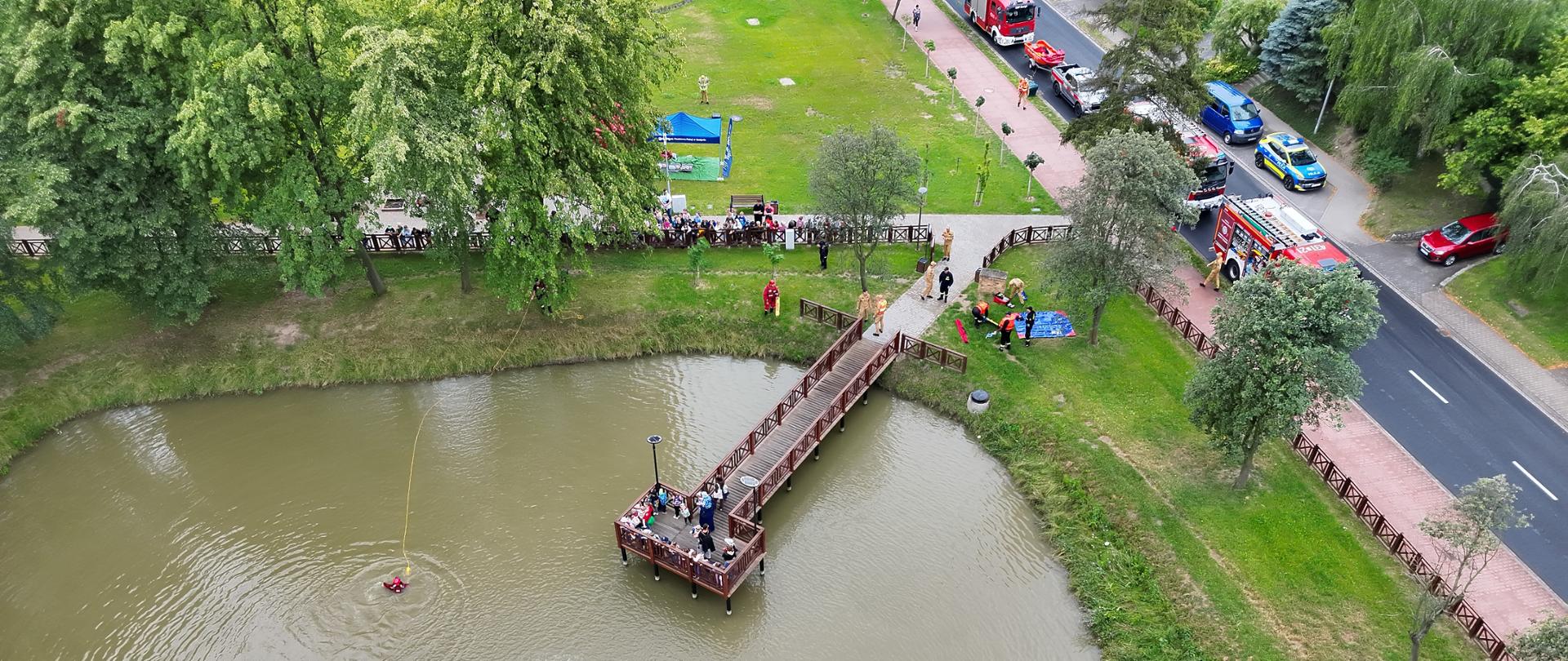 Miejsce ćwiczeń z lotu ptaka. Strażacy przygotowują się do podjęcia osoby poszkodowanej w wody. Przyglądają się temu dzieci w półkolonii i przedszkola. Za nimi na drodze znajdują się wozy strażackie oraz radiowozy policyjne.