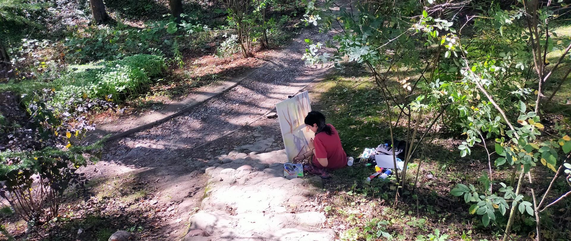 Uczennica siedzi na ziemi i wykonuje pracę malarską. Jest pogodny dzień w Muzeum Wsi Lubelskiej.
