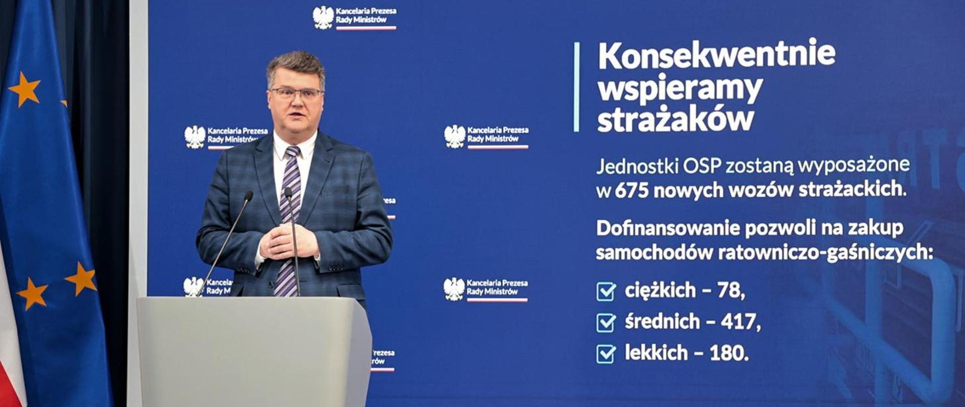 Wiceminister Maciej Wąsik na tle baneru "Konsekwentnie wspieramy strażaków".