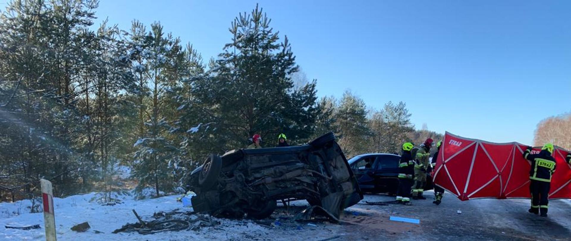 Widać spód rozbitego samochodu, leży na boku poza obrysem drogi, widać również drugi samochód. Na drodze obok rozbitych samochodów strażacy ustawiają czerwony parawan.