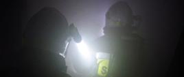 Widok z tyłu. Widoczne sylwetki dwóch strażaków w hełmach z latarkami w ciemnościach w zadymionej piwnicy.