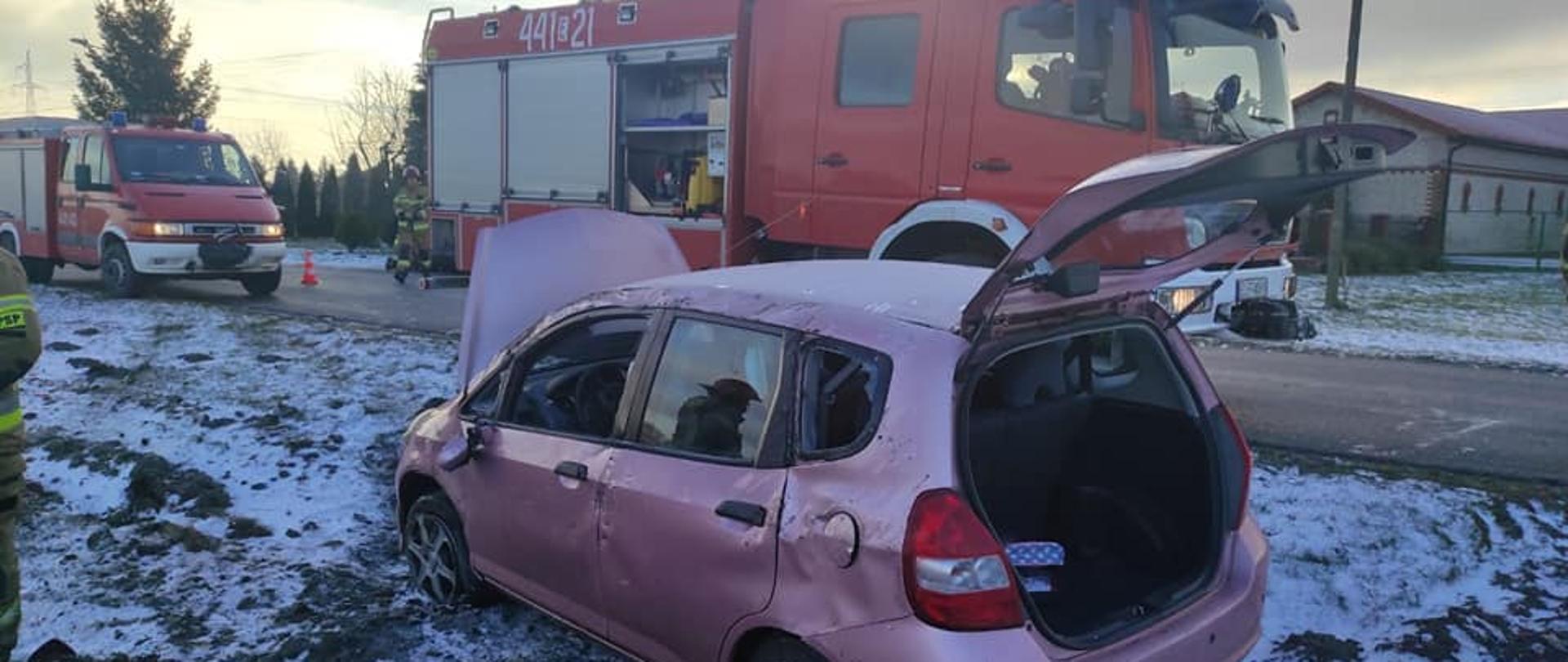 Na zdjęciu widać stojący na poboczu bladoróżowy samochód osobowy z otwartą maską oraz klapa bagażnika. Samochód ma wybite dwie szyby, pogniecioną karoserię oraz urwane lusterko. Obok samochodu na drodze stoi czerwony pojazd straży pożarnej. Samochód ma otwartą jedną skrytkę, w której przewożony jest sprzęt strażacki. Za samochodem straży pożarnej widać drugi mniejszy samochód strażacki. Między samochodami stoi strażak w żółtym stroju i czerwonym hełmem. Na trawie i poboczu jest śnieg. W tle widać zabudowania oraz drzewa i krzewy. Jest widno.