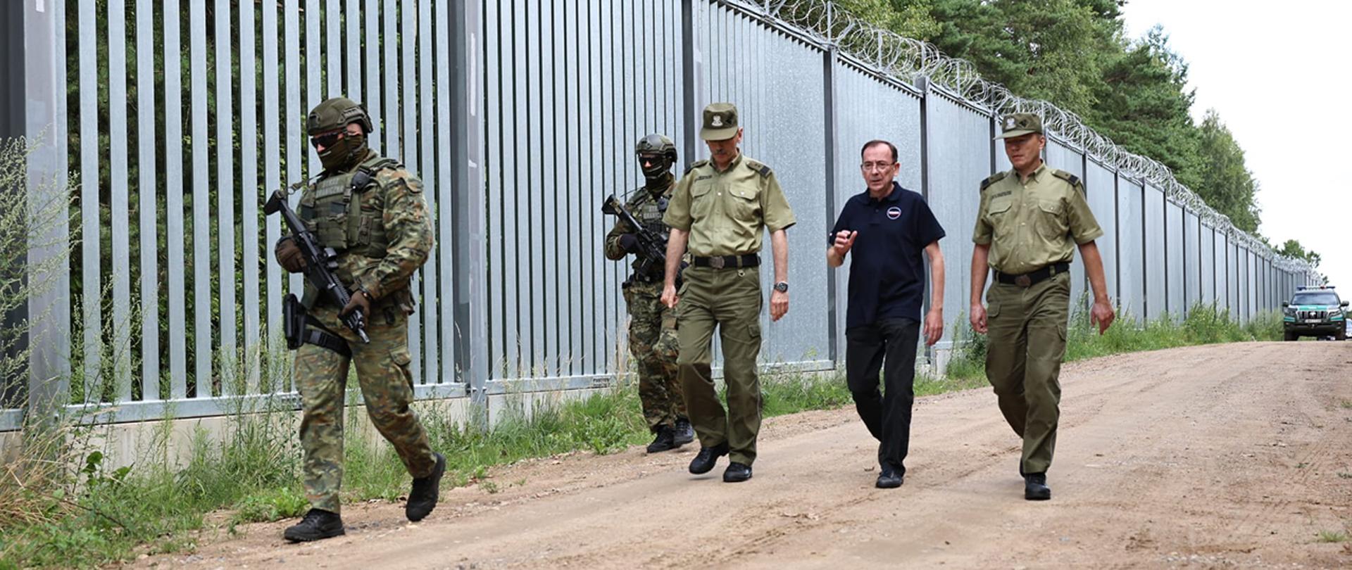 Minister Mariusz Kamiński z komendantem głównym Straży Granicznej i komendantem podlaskiego oddziału Straży Granicznej w Białymstoku idący wzdłuż zapory na granicy polsko-białoruskiej.