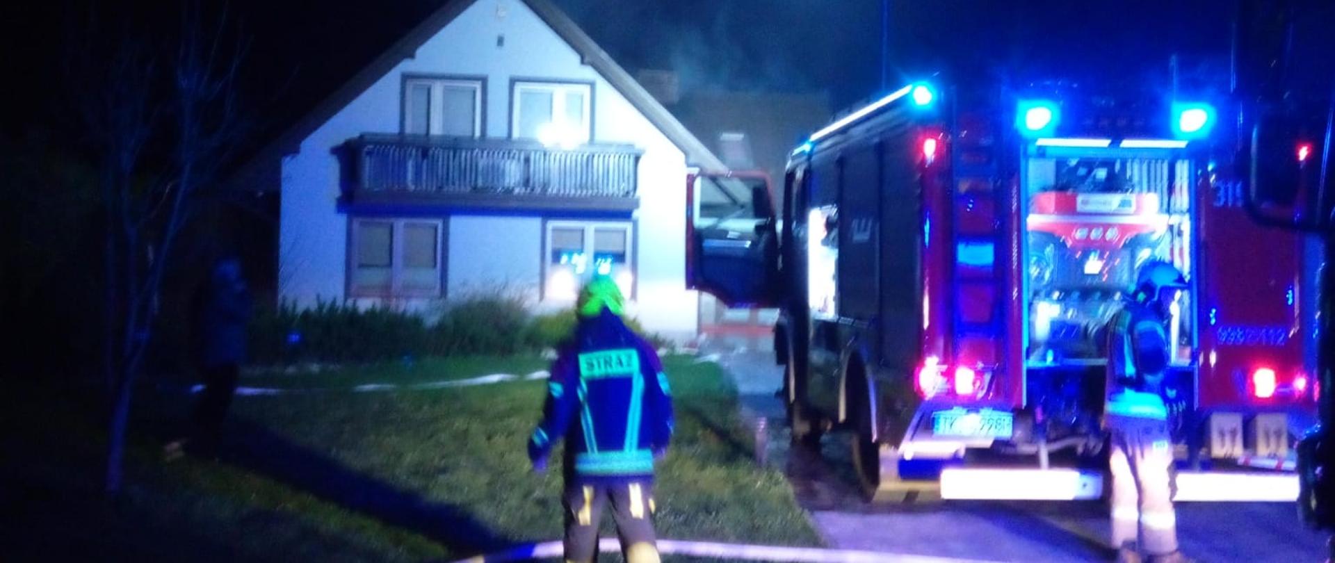 Zdjęcie przedstawia budynek mieszkalny gdzie doszło do pożaru. Na podjeździe stoi samochód gaśniczy a przy nim dwóch strażaków. 