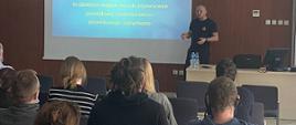 Strażak podczas prowadzonego wykładu z pierwszej pomocy przedmedycznej stoi na tle wyświetlanej prezentacji. Przed nim na krzesłach znajdują się licznie zgromadzeni słuchacze.