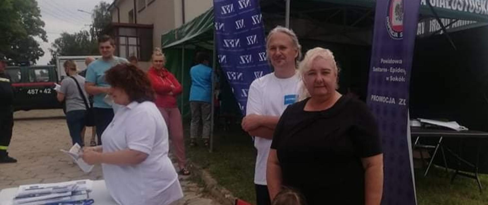 Zdjęcie na drugim planie przedstawia ludzi czekających na szczepienie zgromadzonych przed namiotem typu pawilon oznaczonego napisem "Gmina Dąbrowa Białostocka", w namiocie trwa obecnie szczepienie jednego z nich. Na pierwszym planie, na stole przed namiotem rozłożone są ulotki informacyjne dotyczące szczepienia. Za stołem stoją pracownicy PSSE gotowi do udzielenia wyczerpujących odpowiedzi na ten temat.