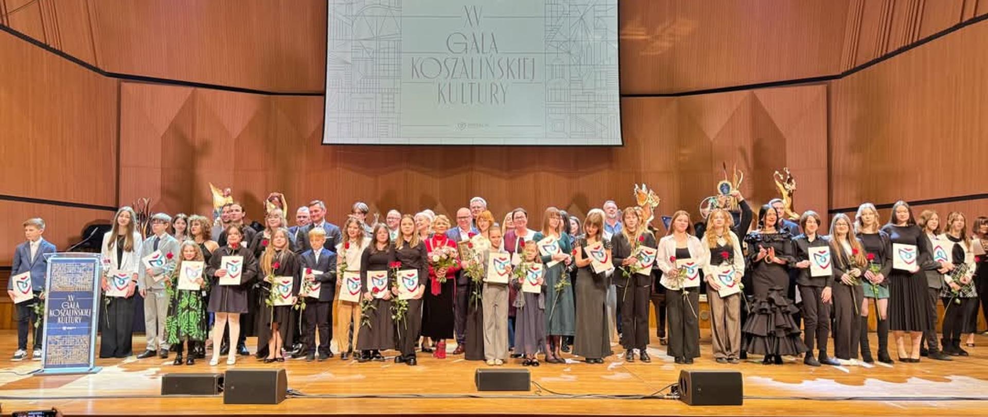 Na scenie w Filharmonii stoją wszyscy nagrodzeni podczas Koszalińskiej Gali Kultury. W tle rozwijany ekran .