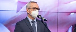 Minister Marek Zagórski podczas konferencji - stoi przed mikrofonem, w białek maseczce na twarzy.