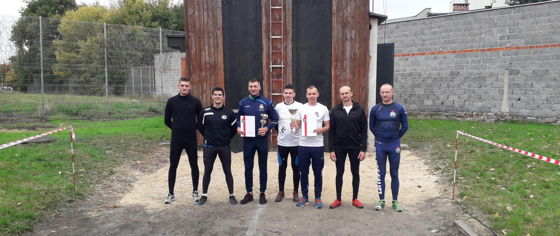 Na zdjęciu widzimy siedmiu mężczyzn w wieku średnim stojących w szeregu i pozujących do zdjęcia. Mężczyźni w szeregu to biorący udział w turnieju, ubrani w kolorowe dresy w rękach trzymają dyplomy oraz złote puchary. Na drugim planie widzimy część konstrukcji wspinalni, ogrodzenie koloru szarego oraz wysoki mur koloru szarego.