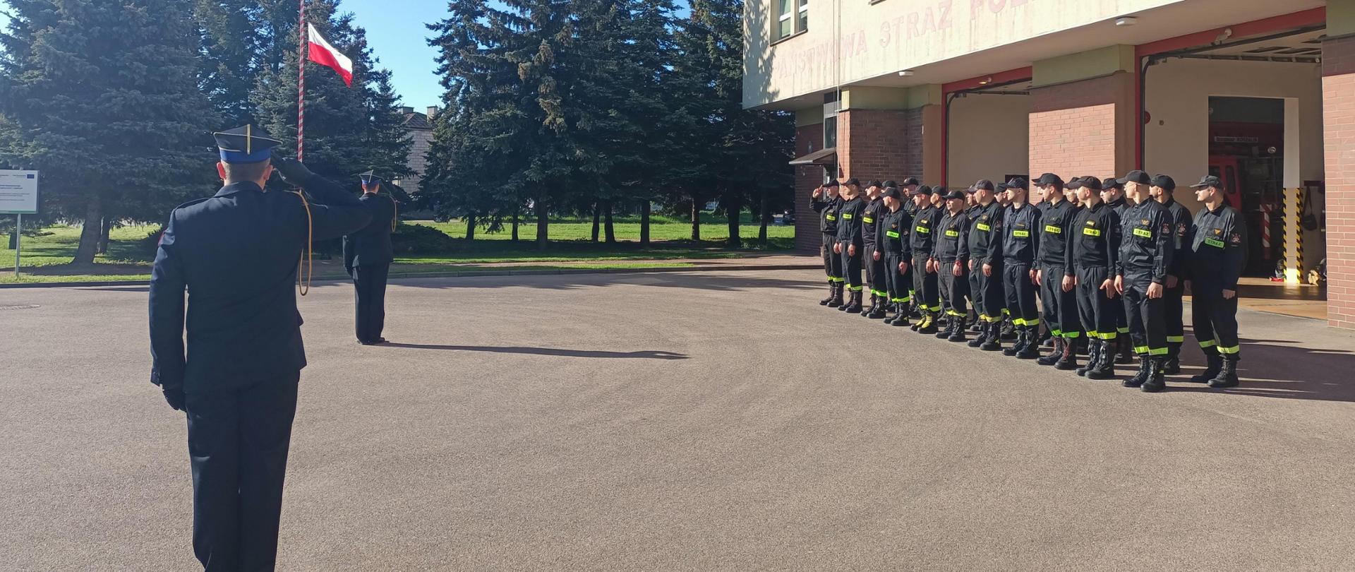 2 strażaków stoi na tle masztu z flagą Rzeczypospolitej Polskiej. Po prawej stronie stoją strażacy podziału bojowego. 