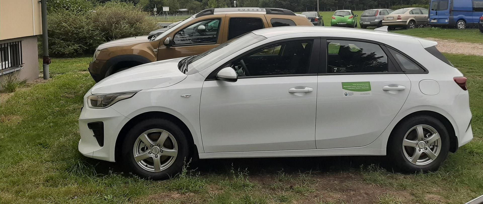 Pojazd koloru białego zaparkowany przed innymi autami i tuż przed budynkiem po lewej stronie. Na tylnych drzwiach widnieje zamieszczona tabliczka z informacją. Za samochodami znajdują się drzewa.