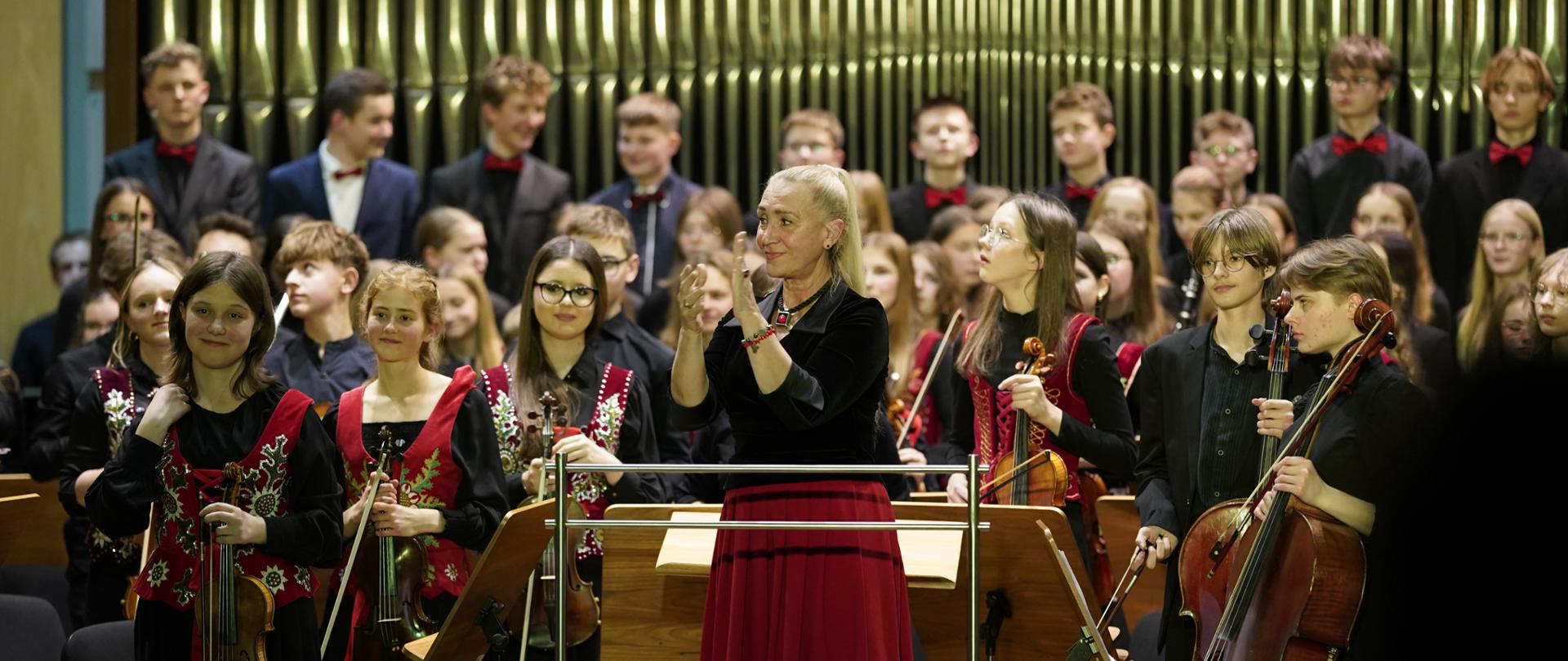 Na zdjęciu widać duży zespół muzyczny złożony z młodzieżowego chóru i orkiestry, wraz z dyrygentką, na scenie w sali koncertowej. Dyrygentka (Centralnie): Starsza kobieta stoi na środku, za pulpitami. Ubrana jest w czarną aksamitną bluzkę i długą, czerwoną spódnicę. Patrzy lekko w prawo i w górę, a jej ręce są uniesione, prawdopodobnie klaszcząc lub dziękując po występie. Ma jasne, długie włosy. Orkiestra (Na pierwszym planie): Dookoła dyrygentki stoją i siedzą członkowie orkiestry. Po lewej stronie widać dziewczęta trzymające skrzypce, ubrane w długie, czarne suknie z czerwono-czarnymi gorsetami z haftami, co nadaje im ludowy charakter. Po prawej stronie stoją osoby grające na wiolonczeli i skrzypcach, ubrane w czarne stroje. Wszyscy spoglądają w kierunku dyrygentki lub publiczności. Chór (W tle): Za orkiestrą, na podwyższeniu, stoją młodzi chórzyści, ubrani głównie na czarno. Chłopcy w tylnych rzędach mają czarne garnitury i czerwone muszki. Wszyscy wydają się być zwróceni do publiczności. Tło: Za chórem dominują duże, pionowe, metalowe piszczałki organowe w ciemnobrązowej oprawie. Pierwszy plan: Na dole zdjęcia widoczne są niewyraźne sylwetki głów publiczności. Zdjęcie przedstawia moment po zakończeniu udanego występu, z dyrygentką w centrum uwagi.