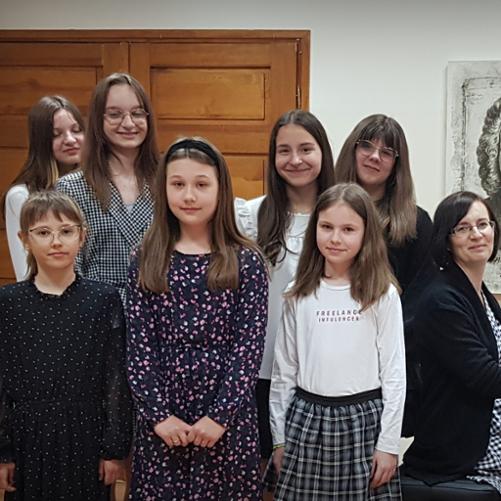 Na zdjęciu widać uczniów Pani Ewy Nowackiej stojących na scenie Auli PSM I st. w Grajewie. Przy fortepianie siedzi nauczycielka Pani Ewa Nowacka. Uczniowie i ich nauczycielka ubrani są w kolory biało, czarno, granatowe. W tle drewniane drzwi.