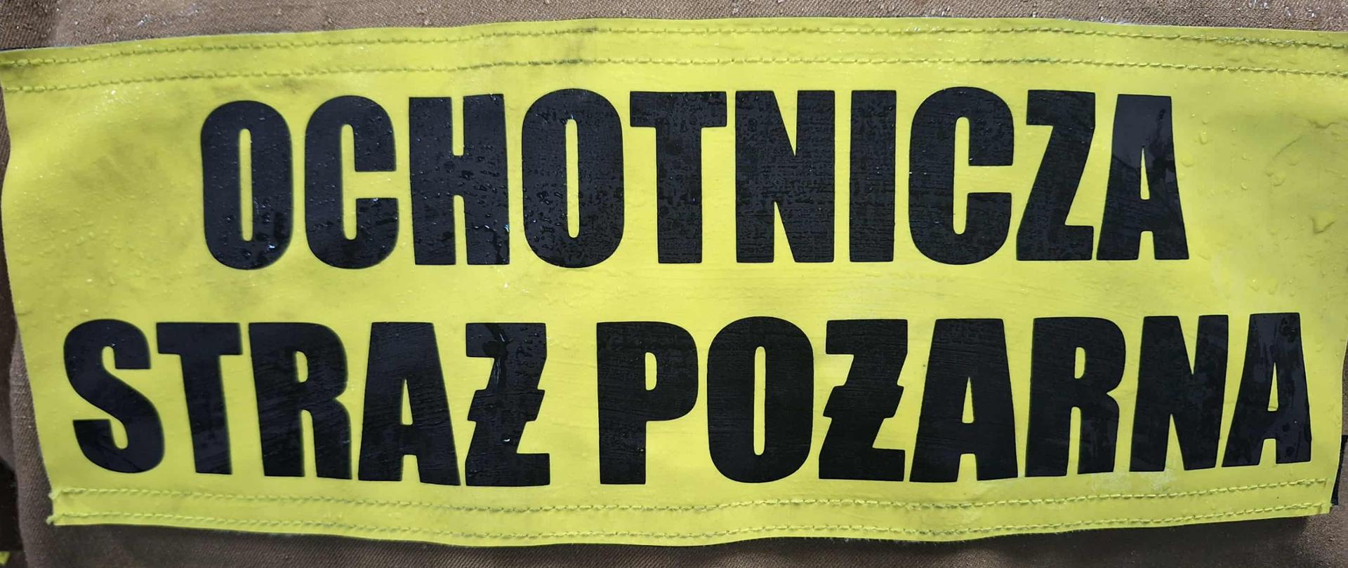 Na zdjęciu widoczny jest fragment ubrania ochronnego strażaka z napisem „OCHOTNICZA STRAŻ POŻARNA”. Napis znajduje się na żółtym odblaskowym tle, a litery są czarne i wyraźne, co zwiększa ich widoczność. Ubranie ma kolor brązowy i jest lekko wilgotne, co może wskazywać na kontakt z wodą lub deszczem. Po bokach widać odblaskowe pasy, typowe dla odzieży ochronnej strażaków. Całość sugeruje, że zdjęcie przedstawia tył kurtki strażaka należącego do Ochotniczej Straży Pożarnej (OSP).
